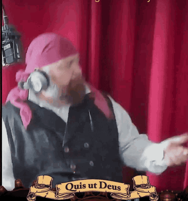a man wearing headphones stands in front of a red curtain and a banner that says " quis ut deus "