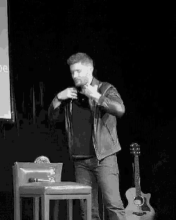 a man in a leather jacket is standing on a stage in front of a microphone and a guitar .