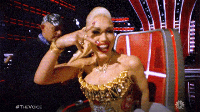 a woman in a gold dress is sitting in a chair with the nbc logo on the bottom