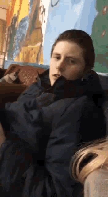 a young man in a blue jacket is sitting in front of a wall with a painting on it .