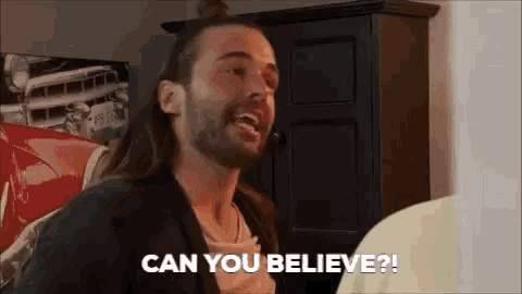 a man with long hair and a beard is talking to another man in a room .