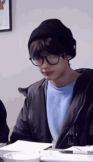 a young man wearing glasses and a beanie is sitting at a table .