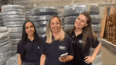 three women are posing for a picture in a warehouse in front of a pile of tires .