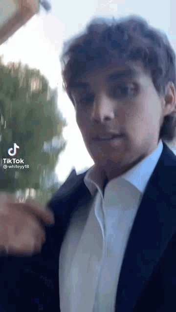 a man in a suit and white shirt is standing in front of a tree .