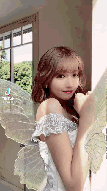 a woman in a white dress with angel wings is standing next to a window .