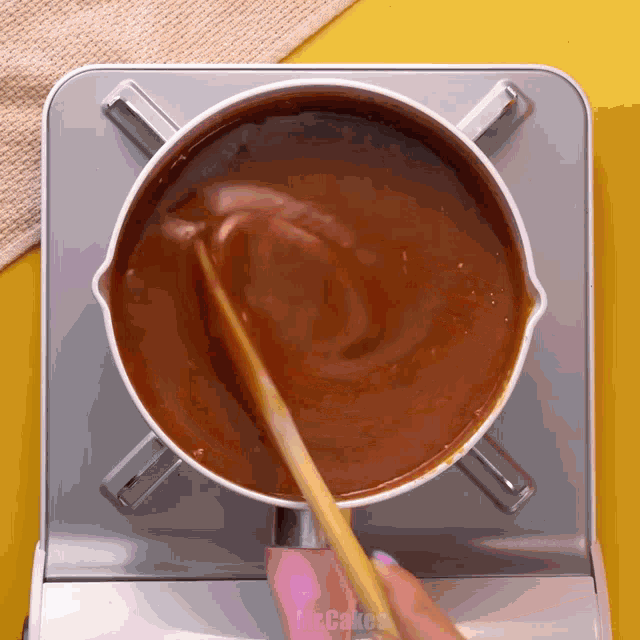 a pot of brown liquid is being stirred with a wooden spoon and the word mrcakes is visible on the stove