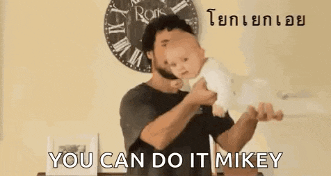 a man is holding a baby in his arms and says `` you can do it mikey '' in front of a clock .