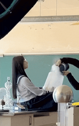 a girl sits on a desk in a classroom while a man holds a container of water