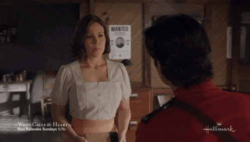 a man in a red uniform is talking to a woman in a white top in front of a wanted poster