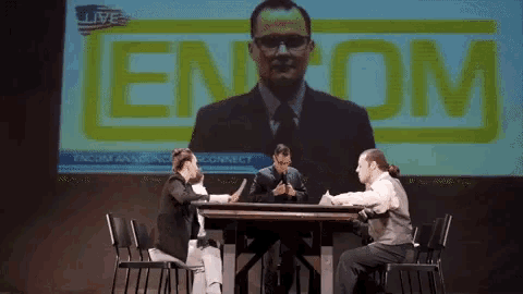 a group of people are sitting at a table in front of a large screen that says encom