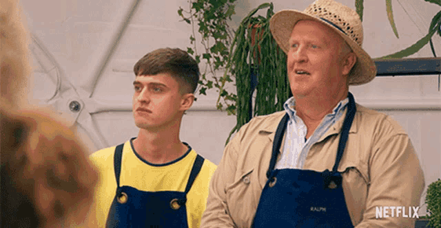 a man wearing overalls and a hat is standing next to another man wearing an apron that says ralph on it