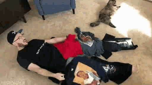 a man is laying on the floor surrounded by clothes and a cat is sitting on the floor .