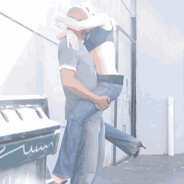 a man is carrying a woman in his arms in front of a green trash can that says ' t.t. '