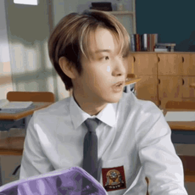 a young man in a white shirt and tie is sitting at a desk in a classroom holding a purple pencil case .