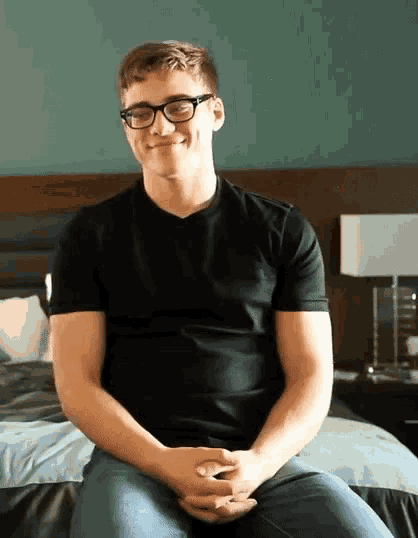 a man wearing glasses and a black shirt sits on a bed with his hands folded
