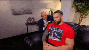 roman reigns is sitting on a couch talking to a man in a suit while wearing a red shirt .