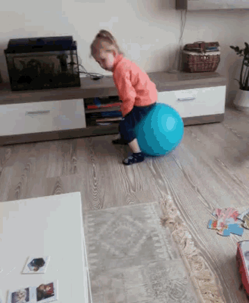 a little girl sitting on a blue ball in a living room