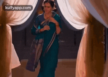 a woman in a blue saree is standing in a room with curtains .