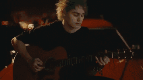 a man with green hair is playing an acoustic guitar in front of a red car .