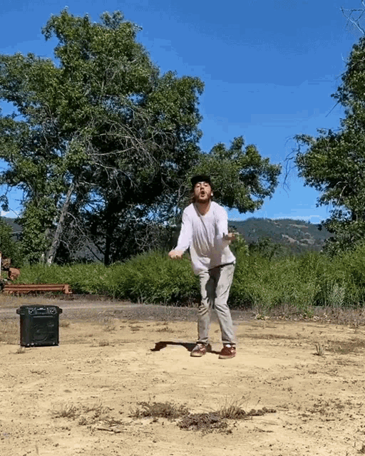 a man in a white shirt and gray pants is standing in a dirt field with trees in the background