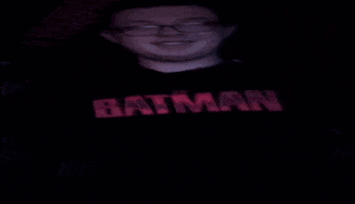 a young man wearing glasses and a black batman shirt is making a funny face .