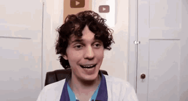 a young man with curly hair is smiling while sitting in a chair in a room .