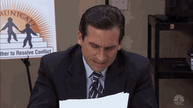 a man in a suit and tie sits in front of a sign that says communicate