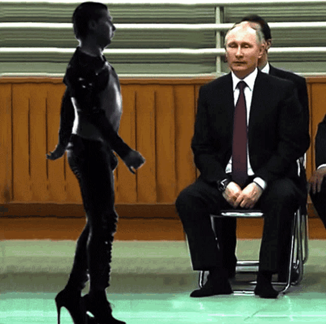 a man in a suit and tie sits in a chair while a woman stands in front of him