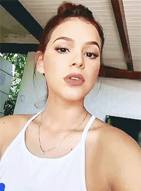 a woman wearing a white tank top and a gold necklace takes a selfie