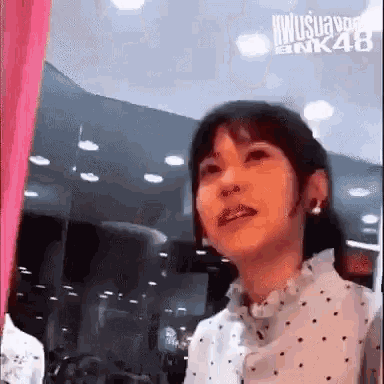a woman in a white polka dot shirt is standing in front of a pink curtain in a room .