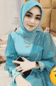 a woman in a blue dress and hijab is holding a cell phone in her hand .