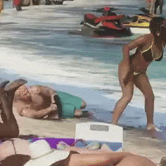 a woman in a bikini is standing on a beach next to a man laying on the sand .