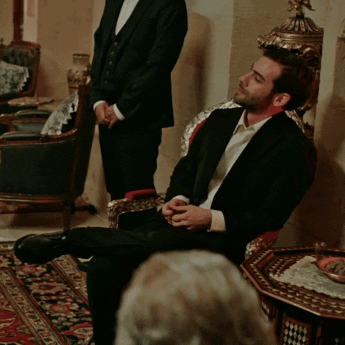 a man in a tuxedo is sitting in a chair with his legs crossed