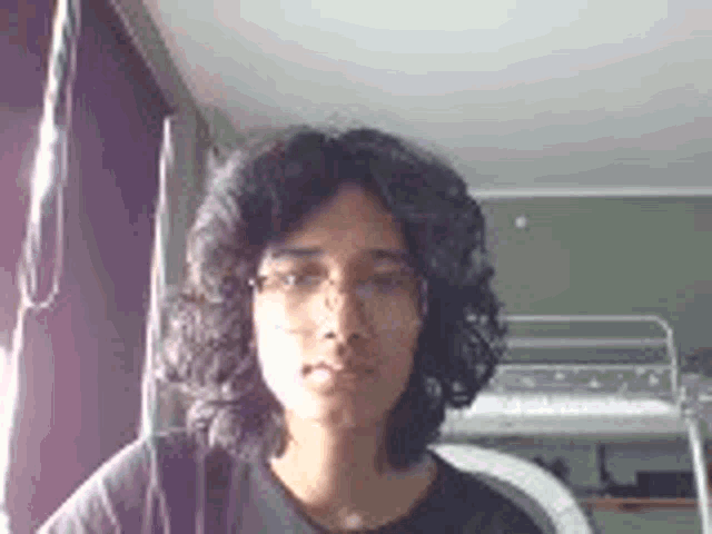 a young man with long curly hair and glasses is looking at the camera in a bedroom .
