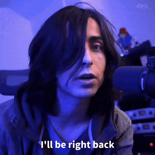 a man with long hair is talking into a microphone and saying i 'll be right back