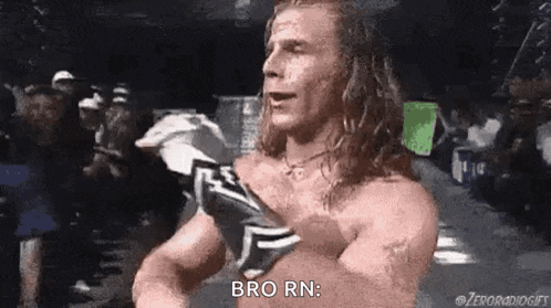 a shirtless wrestler with long hair is standing in front of a crowd and talking .