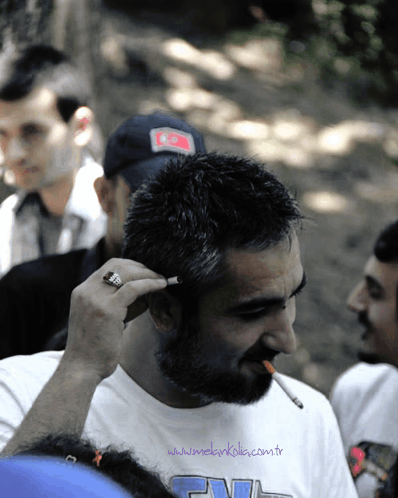 a man with a beard is smoking a cigarette and wearing a white shirt that says www.mekarkola.com.tr