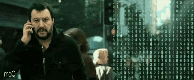 a man with a beard is talking on a cell phone in front of a screen that says mq