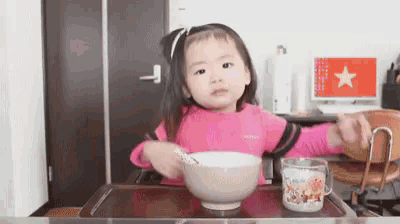 a little girl is sitting at a table with a bowl of food and a cup with an anpanman design on it