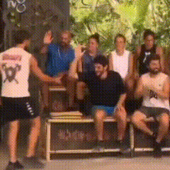 a group of people are standing and sitting on a bench with one man wearing a white shirt with a x on it .