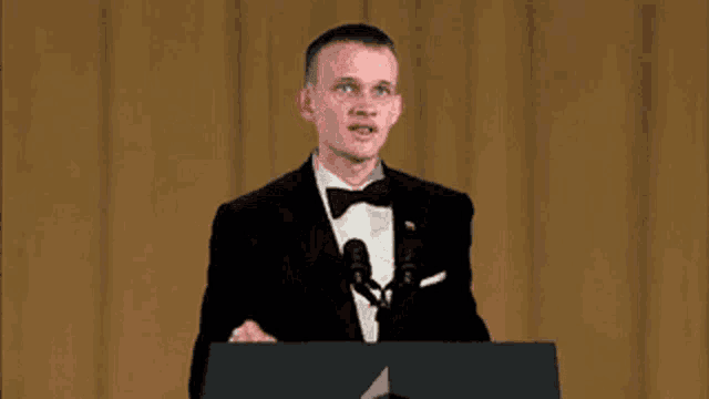 a man in a tuxedo stands at a podium speaking into a microphone