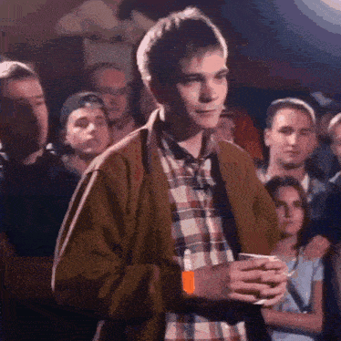 a young man in a plaid shirt is standing in a crowd of people holding a cup .