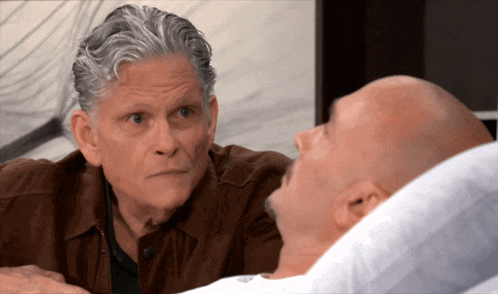 a bald man is laying in a hospital bed while another man looks on