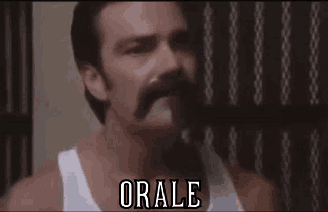 a man with a mustache is wearing a white tank top and saying orale .