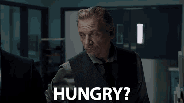 a man in a vest and tie says hungry in a dark room
