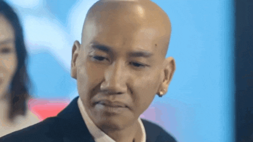 a bald man in a suit and tie is looking at the camera with a woman in the background .