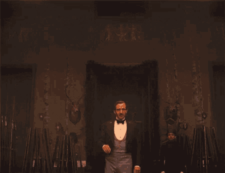 a man in a tuxedo and bow tie stands in a dark room surrounded by guns