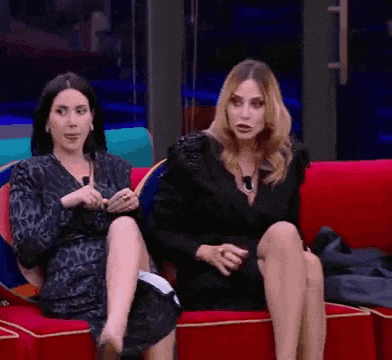 two women are sitting on a red couch with their legs crossed and talking to each other .