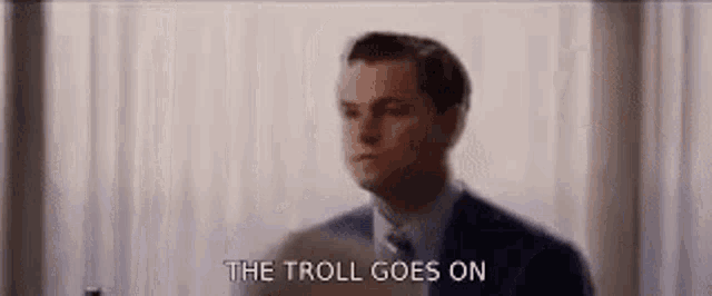 a man in a suit and tie is standing in front of a window and says the troll goes on .