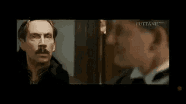 a man with a mustache is talking to another man in a room .